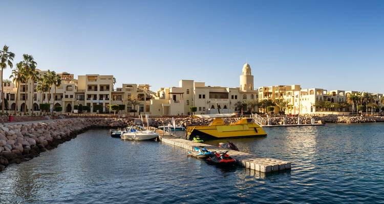 Sonnige Marina mit modernen beigen Gebäuden, Booten und einem weißen Minarett neben ruhigem blauen Wasser.