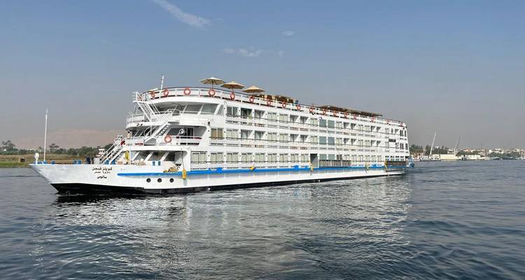 Un bateau de croisière blanc sur le Nil, à plusieurs ponts, glisse sur des eaux calmes sous un ciel brumeux.