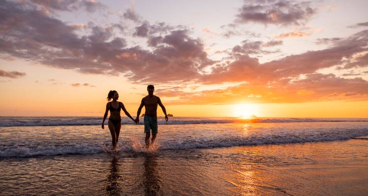 Silhouetgevormd stel dat hand in hand door het zachte golfgebeuren op een tropisch strand bij zonsondergang loopt.