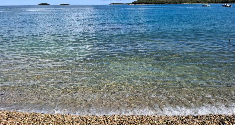 De petites vaguettes caressent une plage de galets avec une mer bleu calme et des îlots lointains