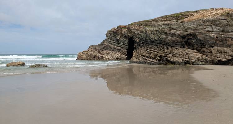 Plage de sable battue par les vagues avec une grande formation rocheuse de grotte se reflétant sur le sable mouillé