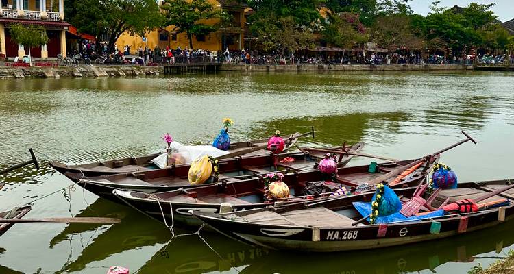 Holzerne Sampan-Boote, die mit farbenfrohen Papierlaternen geschmückt sind, treiben auf einem ruhigen Fluss vor gelben Gebäuden.