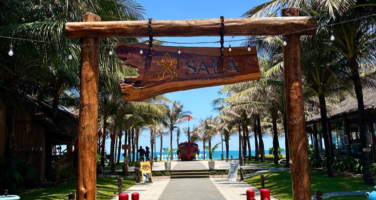 Holzernes Eingangschild des SALA Tuy Hoa Beach Club, das sich zwischen einem palmengefaessten Weg zum Meer befindet.