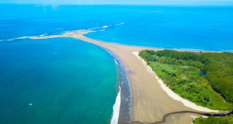 Vista aérea de la barra de arena en forma de cola de ballena extendiéndose hacia el océano azul intenso junto a la selva exuberante.