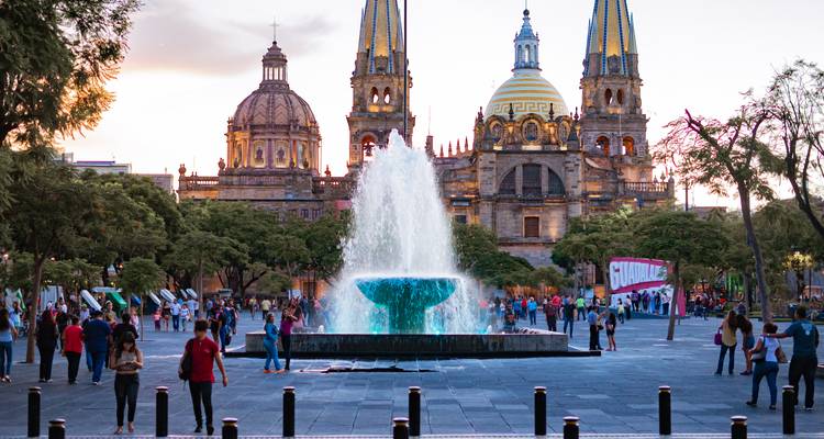 Place animée avec fontaine centrale encadrée par la Cathédrale de Guadalajara aux deux tours jumelles au crépuscule.