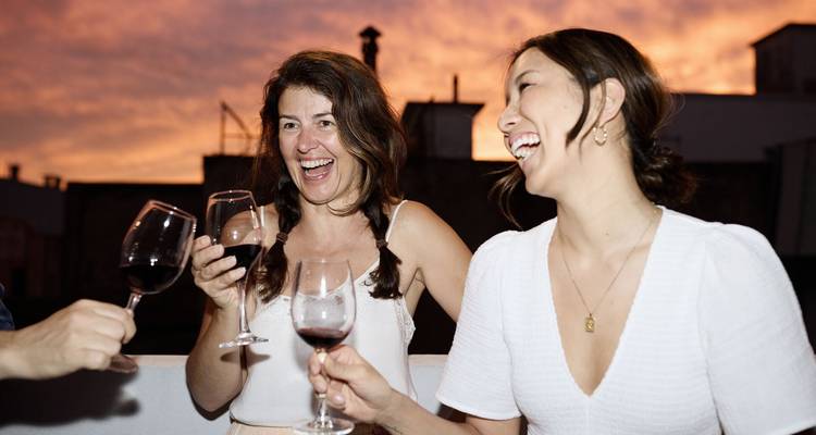 Amigos se ríen y brindan vino tinto en una terraza en la azotea contra un cielo de atardecer ardiente.