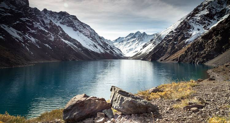 Un tranquilo lago glacial refleja las nevadas cimas de los Andes bajo un cielo pálido.