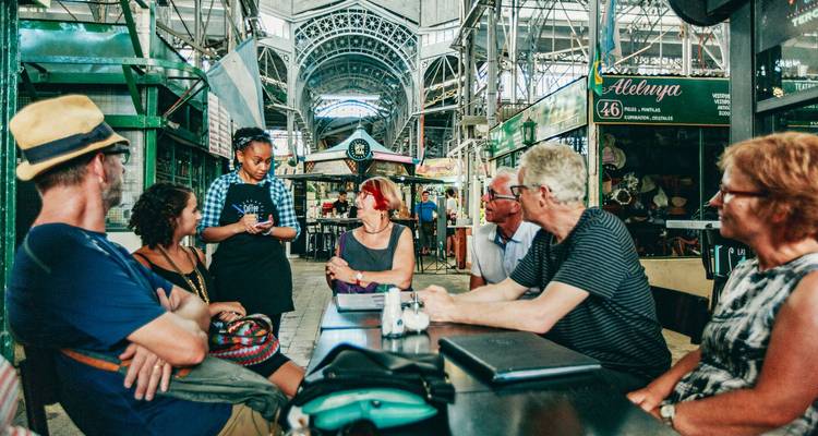 Los viajeros se sientan en una mesa larga dentro de un mercado cubierto lleno de vida mientras una mesera toma los pedidos.