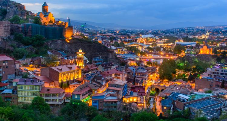 Las luces nocturnas iluminando la ciudad vieja de Tbilisi y las colinas bajo un cielo azul cada vez más oscuro.
