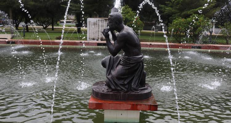 Bronze statue of a thoughtful figure surrounded by fountain jets in a tranquil park pond.