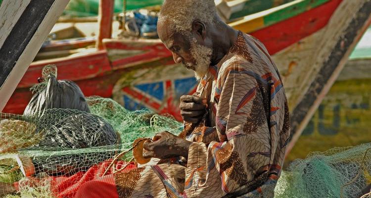 Elderly fisherman repairs green nets beside colorful wooden boats in a coastal village.