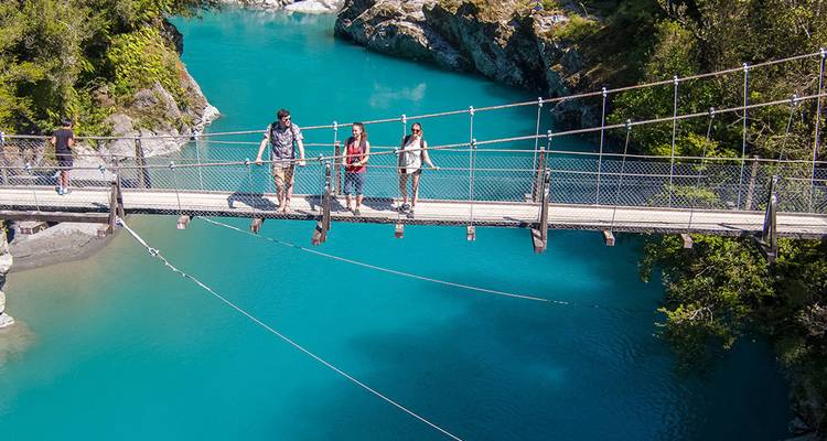 Des voyageurs traversant un pont suspendu étroit enjambant une rivière d'un bleu frappant entourée de montagnes boisées.