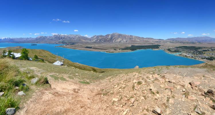 Panorama depuis un point de vue surélevé du magnifique lac turquoise de Tekapo, bordé par des montagnes et des collines arides.