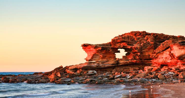 Raue rote Felsformation an der Küste, die bei Sonnenuntergang leuchtet, während Wellen den sandigen Strand darunter umspülen.