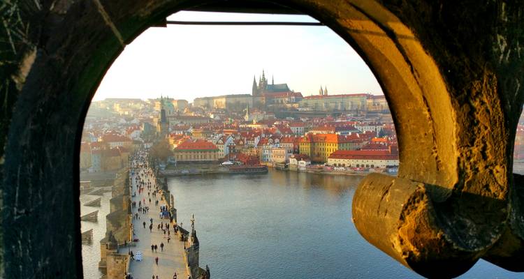 Stone window arch frames a golden view of Prague Castle and Charles Bridge over the river.