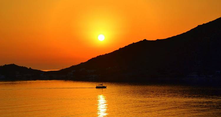 Hermosa puesta de sol anaranjada sobre el mar tranquilo con la silueta de una colina y una pequeña barca cruzando el agua brillante.