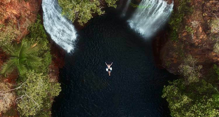 Drohnenaufnahme eines Schwimmers, der zwischen zwei Wasserfällen schwebt, die in einen dunklen Pool mit üppig grüner Umgebung stürzen.