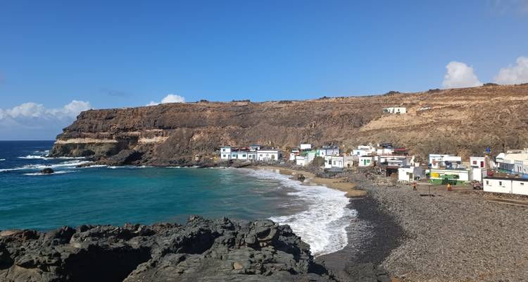Petit village côtier de maisons blanches bordant une baie de galets avec une eau turquoise et des falaises escarpées.