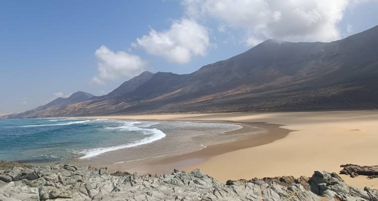 Large plage de sable doré bordée par une mer bleue cristalline et une chaîne de montagnes spectaculaire sous un ciel lumineux.
