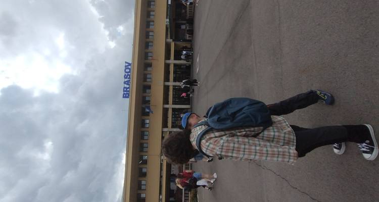 Des personnes traversant la cour d'un bâtiment avec une grande enseigne BRASOV sous un ciel nuageux.