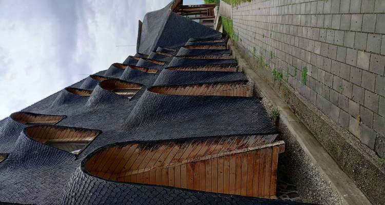 Bâtiment moderne en bois avec façade en bardeaux ondulants le long d'une promenade pavée sous un ciel gris.