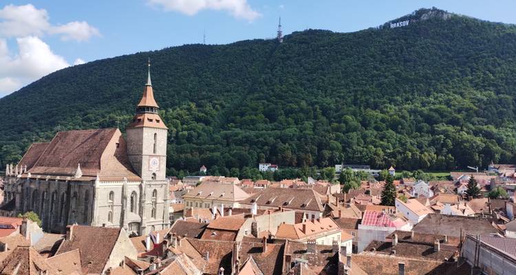 Vue surélevée sur les toits de tuiles rouges de la vieille ville de Brasov avec l'Église Noire et les collines boisées en arrière-plan.