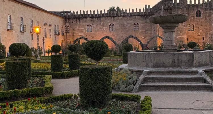 Scène de jardin en soirée avec topiaires taillés et murs en pierre historiques doucement illuminés par des lampes.