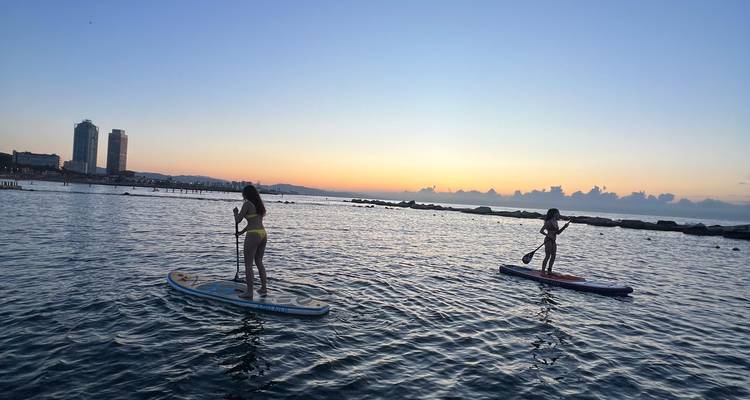 Zwei Paddleboarder, die bei Tagesanbruch über sanfte Wellen gleiten, mit Stadtsilhouette im Hintergrund