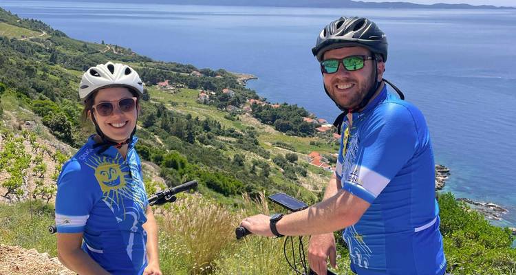 Des cyclistes souriants font une pause au sommet d'une colline avec des vues panoramiques sur la mer et des villages côtiers en contrebas.