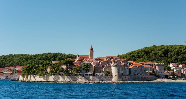 La ville de Korčula aux murs de pierre avec ses toits rouges vue depuis l'eau face à une colline boisée.