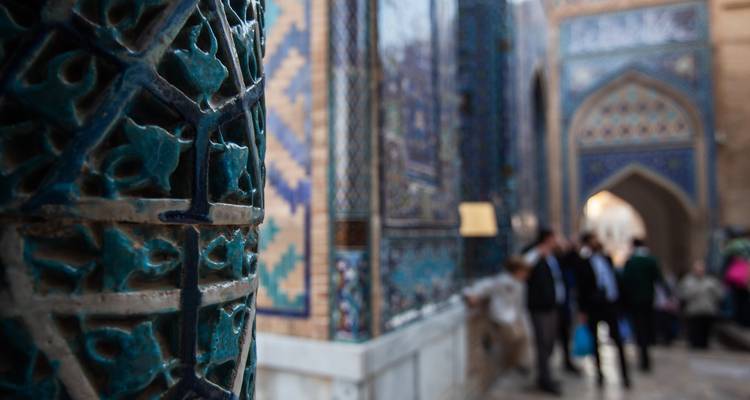 Artistic close-up of turquoise glazed tilework with blurred visitors in a Samarkand mausoleum corridor.