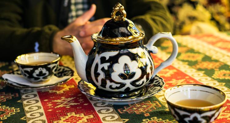 Decorative Uzbek teapot and cups sit on a patterned tablecloth with hands folded in the background.