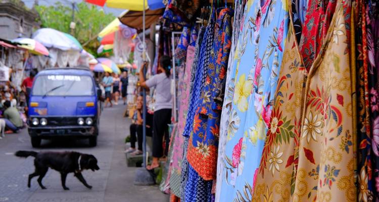 Les étals de tissus colorés bordant un marché de rue balinais animé avec des habitants, des touristes, un chien qui passe et un minibus bleu.