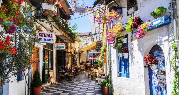 Callejón lleno de flores con cafés y pavimento a cuadros en un soleado pueblo mediterráneo