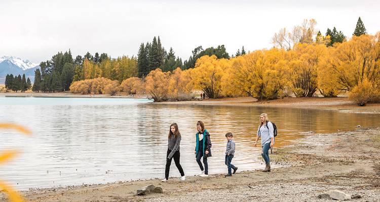 Une famille se promène le long du rivage d'un lac calme bordé d'arbres d'automne dorés et de pics enneigés au loin.