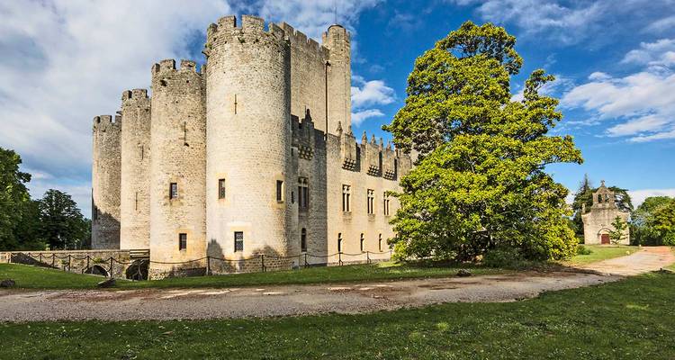Château de pierre médiéval aux tours crénelées situé au milieu de jardins luxuriants sous un ciel partiellement nuageux dans le sud-ouest de la France.
