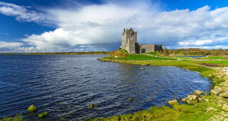 El solitario Castillo de Dunguaire medieval en una lengua de tierra cubierta de hierba que se adentra en un lago sereno bajo cielos azules vibrantes
