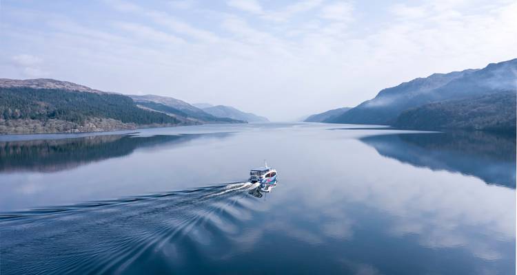 Boat cruising across the glassy waters of a Scottish loch surrounded by misty hills