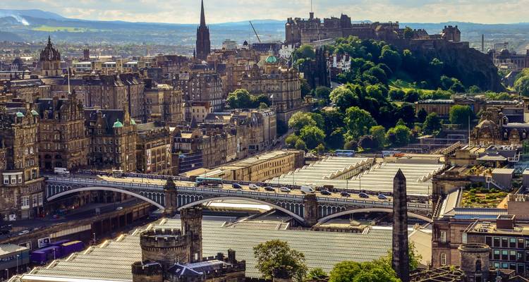 Panoramic aerial of Edinburgh with Waverley Bridge, Scott Monument and Edinburgh Castle atop lush hills.