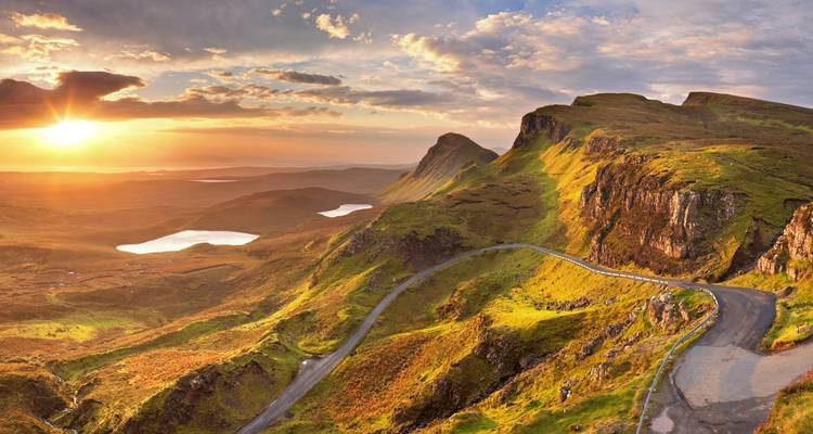 Dramatic winding road through rugged green cliffs and lochs under a glowing sunrise on the Isle of Skye.