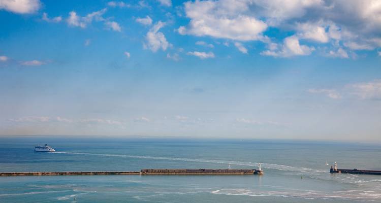 Le traversier s'éloigne d'une longue digue dans la mer ouverte sous un ciel bleu éclatant avec des nuages épars.