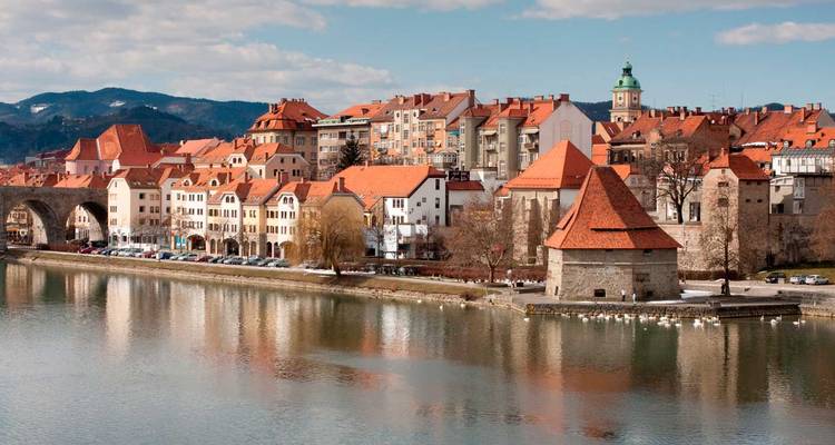 Skyline riverain de Maribor avec des toits en tuiles rouges et les reflets du fleuve Drava par une journée dégagée.