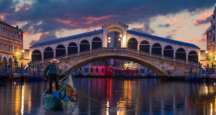 Gondolier lenkt Gondel unter der beleuchteten Rialto-Brücke, die sich im Großkanal von Venedig in der Dämmerung spiegelt