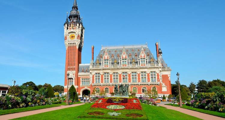 L'hôtel de ville de Calais avec sa haute tour d'horloge et son jardin de fleurs colorées sous un ciel bleu dégagé.