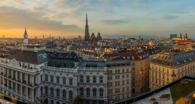 Large panorama sur les toits de Vienne dominée par la haute flèche de la cathédrale Saint-Étienne au coucher du soleil avec des collines lointaines à l'horizon.