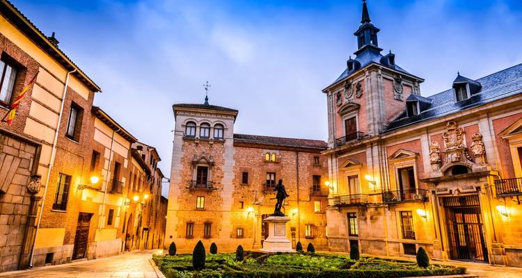 Historischer Platz in Madrids Altstadt mit Laternenbeleuchtung, einer zentralen Statue und eleganten Fassaden in der Dämmerung
