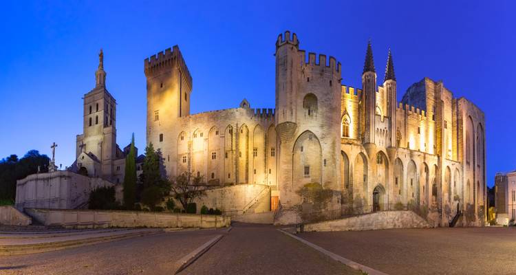 Panorama crépusculaire du Palais des Papes, forteresse-palais illuminé contre un ciel bleu profond à Avignon.