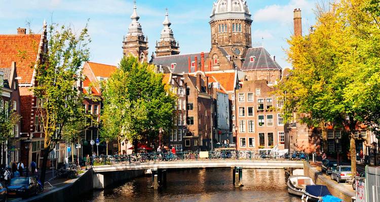 Scène du canal d'Amsterdam avec des bicyclettes, des arbres feuillus et les dômes de la Basilique Saint-Nicolas derrière un petit pont.