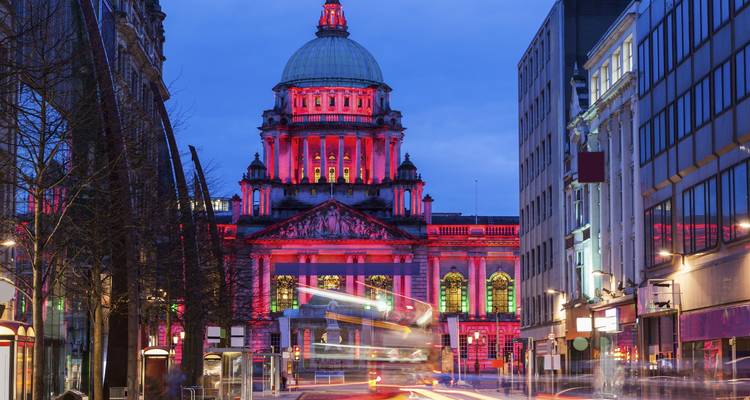 Das Belfast City Hall, das in der Abenddämmerung in leuchtenden Farben erleuchtet ist, mit Lichtspuren von vorbeifahrenden Bussen auf der Straße.