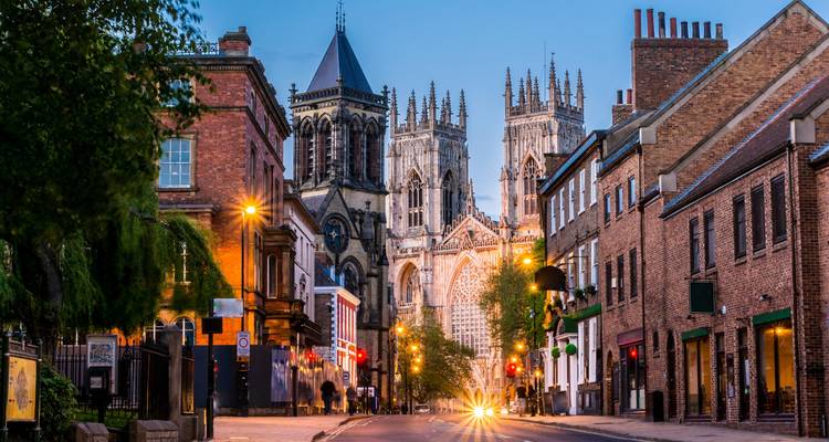 Dämmerungsszene einer Straße in York mit Blick auf die beleuchtete York Minster hinter Backsteinreihenhaüsern.